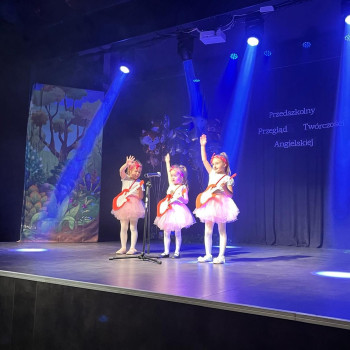 Trzy dziewczynki z gitarami w jasnych strojach na scenie oświetlonej niebieskim światłem